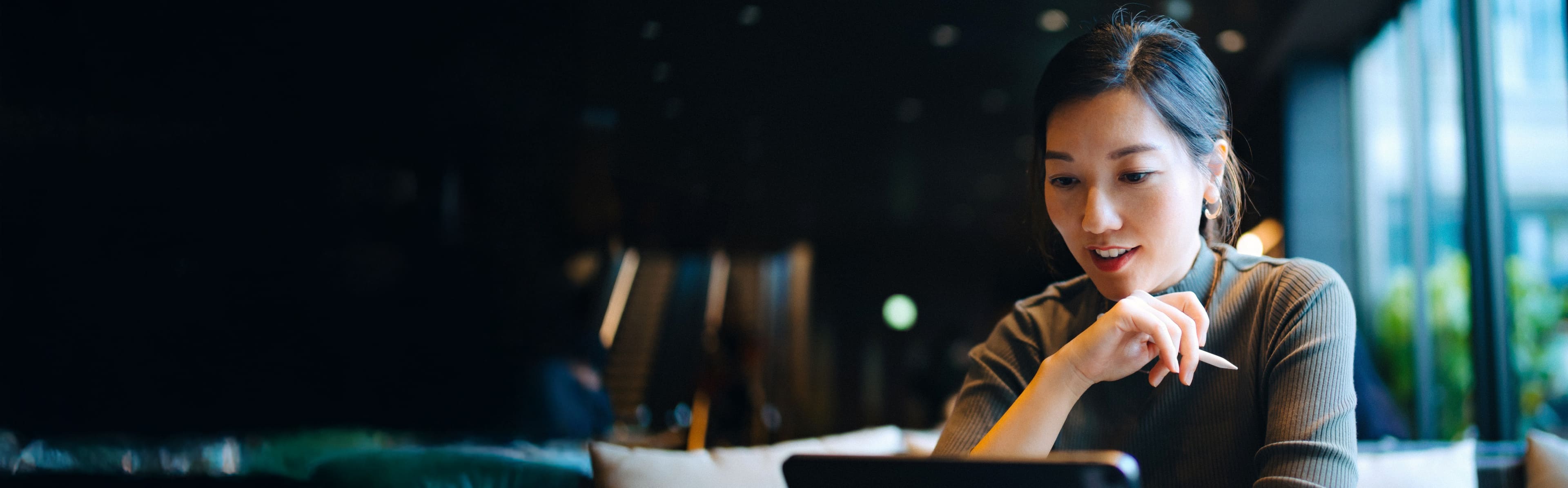 Person sitting at a table in a dimly lit room, looking at a laptop screen with hand resting on chin.