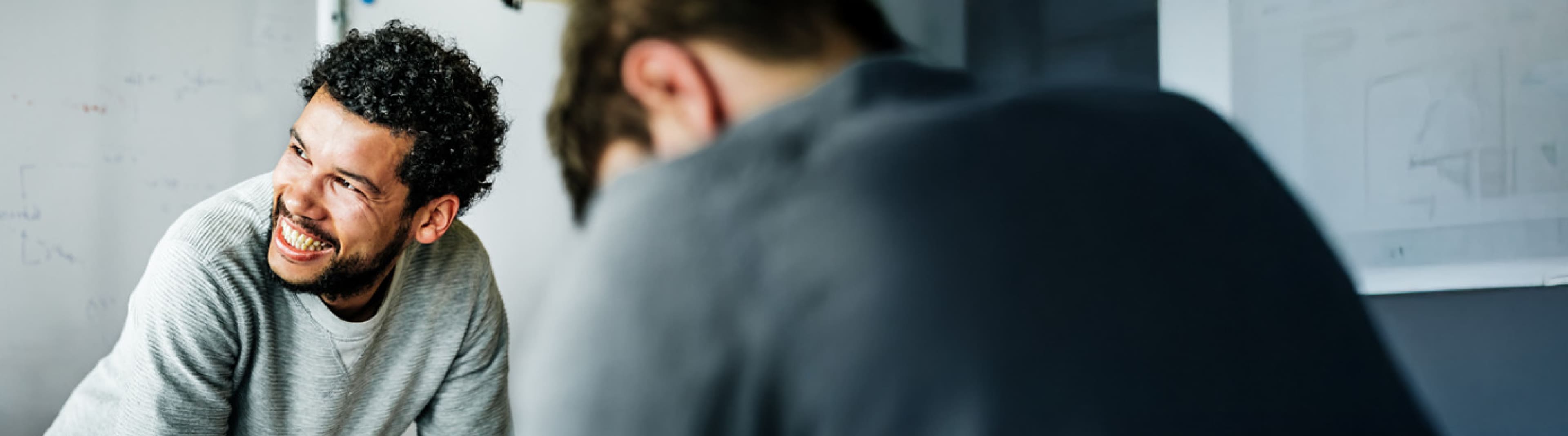 Two individuals engaged in discussion; one facing the camera with a blurred face and wearing a light grey sweater, the other with back to the camera in a dark grey shirt, with indistinct drawings on the wall behind them.