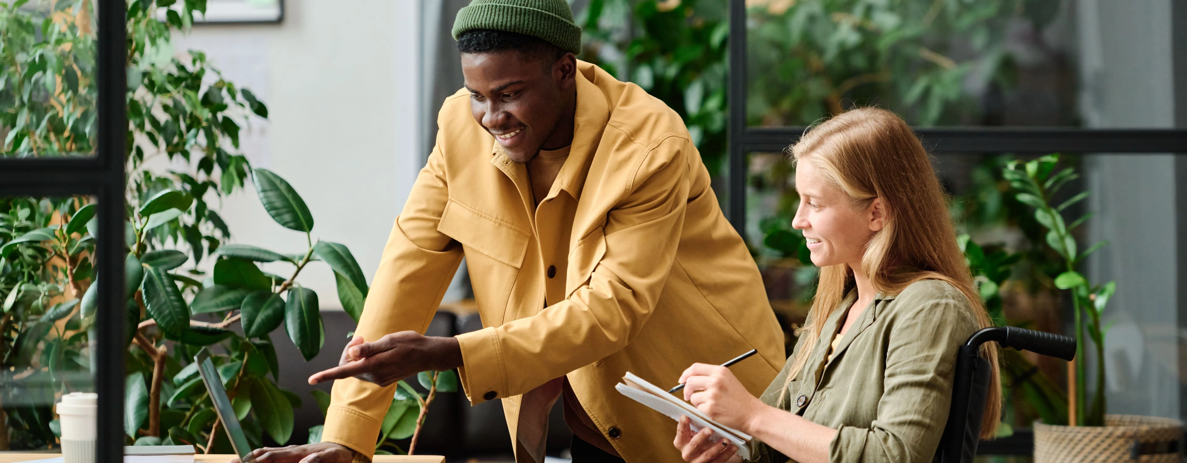 Two people in an indoor setting with plants and a wooden table. One person in a wheelchair holds a notebook and pen, while the other stands beside them pointing at a laptop screen. Faces are blurred for privacy.