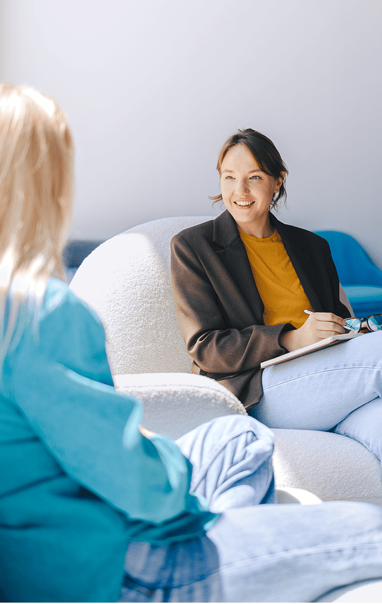Two people sitting and facing each other in conversation. One wears a blue top with blonde hair, and the other wears a brown jacket over a yellow shirt, holding a notepad and pen. Background includes indoor furniture like chairs or couches.