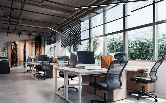 Modern office space with multiple workstations, each equipped with a desk, office chair, and computer monitor. Natural light streams through large windows, with indoor plants and decorative elements along the back wall. Exposed ceiling pipes and industrial lighting complete the contemporary design.